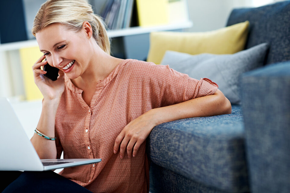 Eine Frau arbeitet auf einem Laptop, während sie vor einem Sofa sitzt und ein Handy ans Ohr hält.
