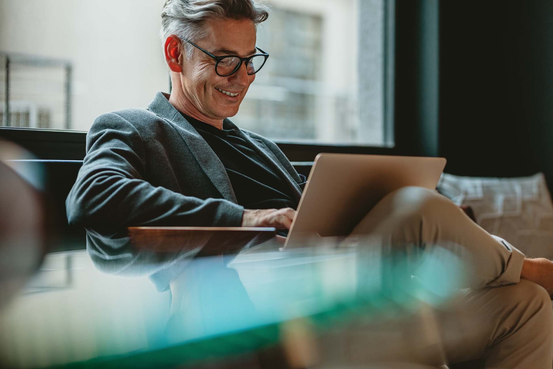 Auf einer Couch sitzt ein Mann mit Brille und nutzt einen Laptop.