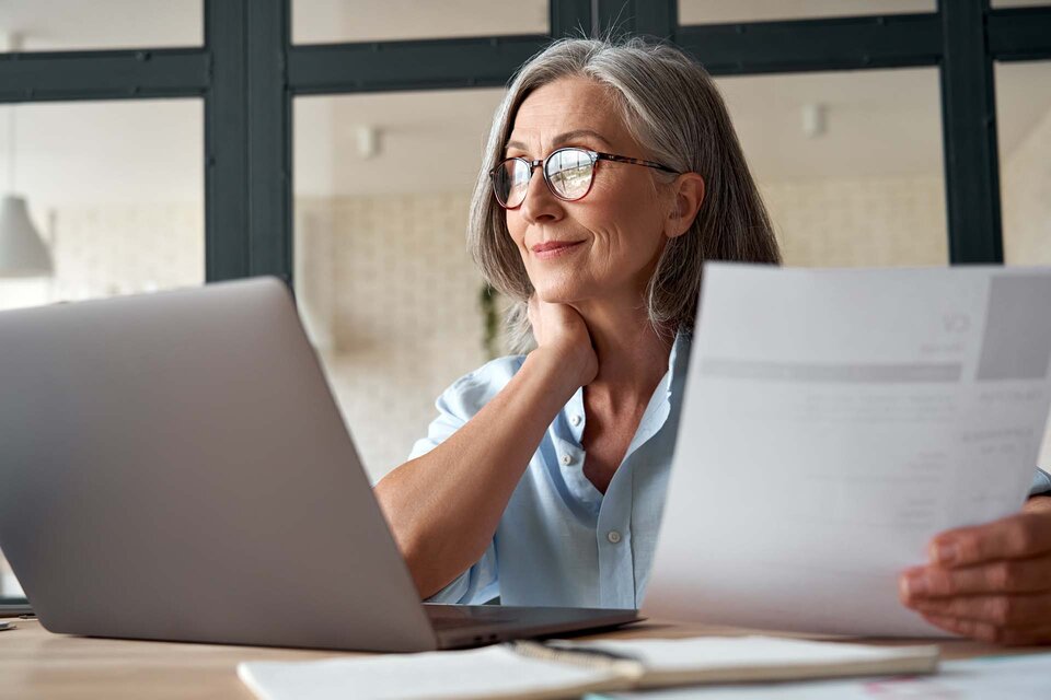 Eine ältere Frau mit Brille sitzt an einem Schreibtisch und arbeitet an einem Laptop.