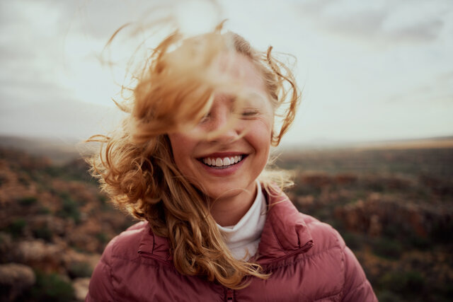 Eine Frau, deren Haare um ihren Kopf im Wind wehen, schließt die Augen und lächelt.