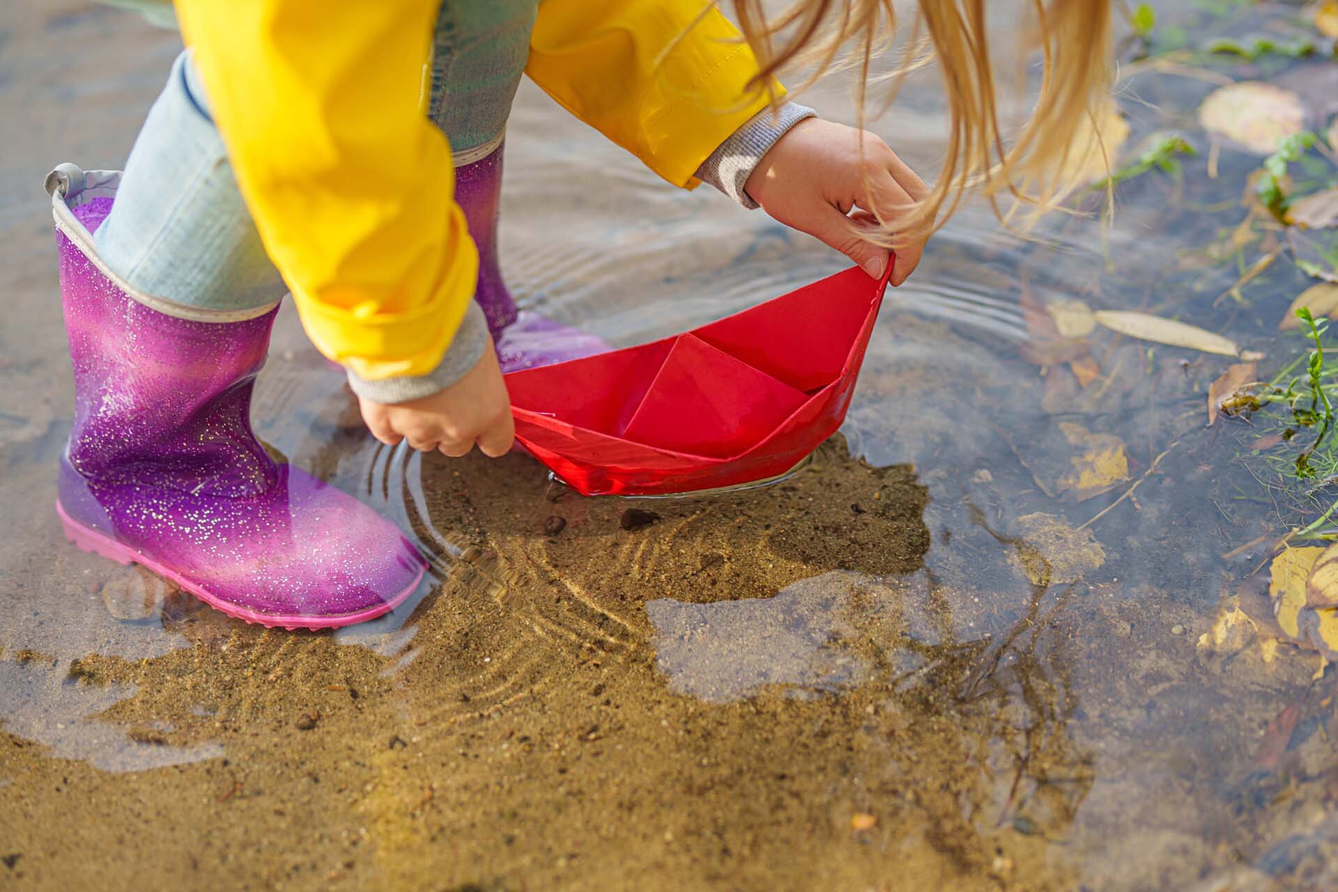 Ein Kind in einem gelben Regenmantel und lila Stiefeln setzt ein rotes Papierboot in eine Pfütze.