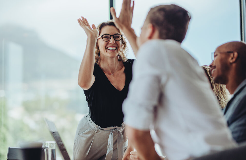 Eine Gruppe von Menschen sitzen in einem Büro, während eine Frau begeistert die Hand hebt und einen High Five gibt.