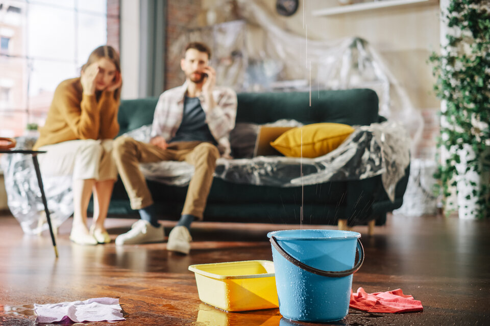 Leitungswasserschaden in der Wohnung – welche Versicherung zahlt wann? | © Getty Images/iStockphoto Das Bild zeigt ein junges Paar, das in einem Wohnzimmer sitzt, in dem ein Leitungswasserschaden aufgetreten ist. | © Getty Images/iStockphoto