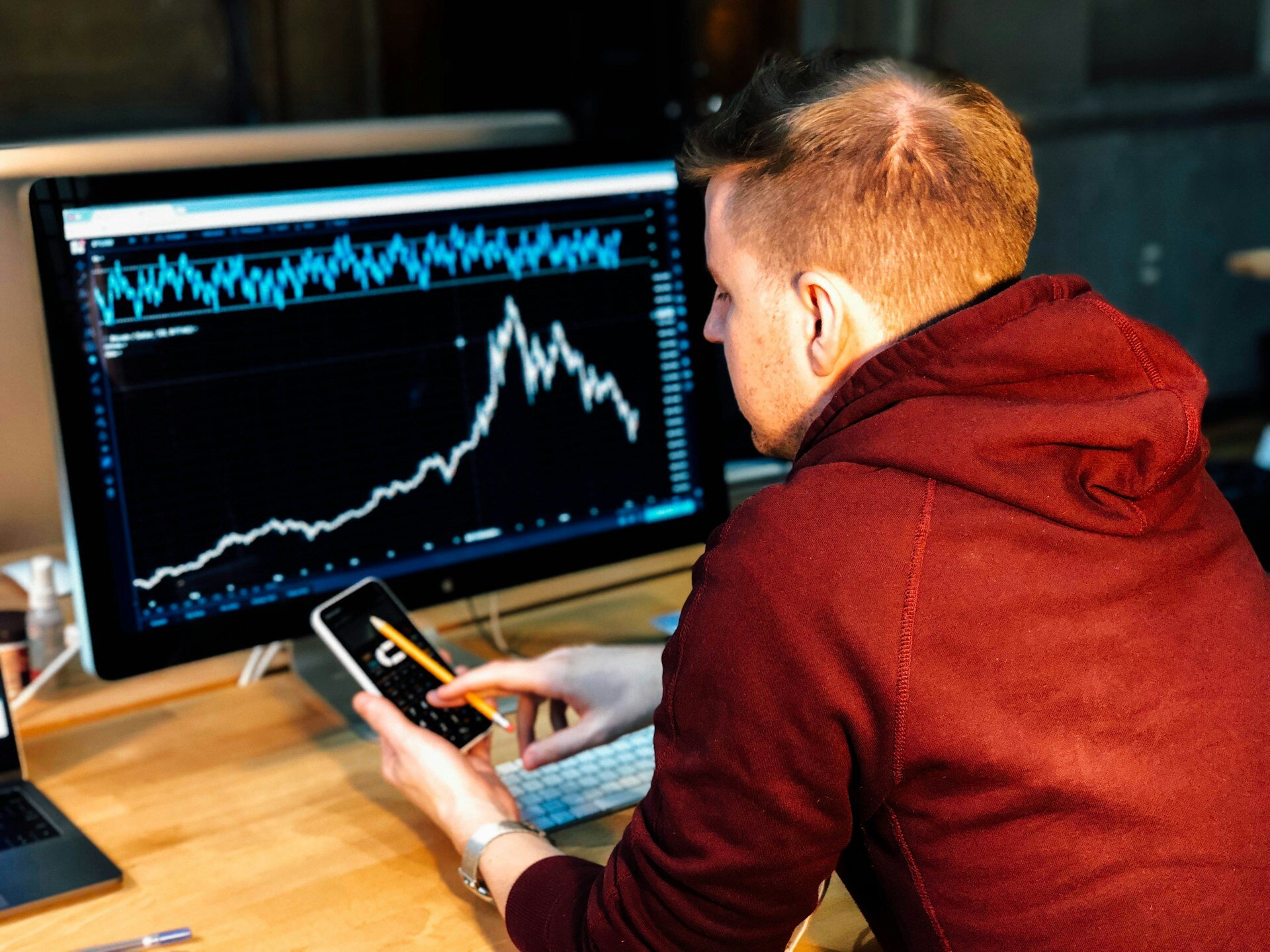Mann hält schwarzes Smartphone vor Computer und schaut sich Aktienkurse auf beiden Endgeräten an. | © Chris Liverani /Unsplash.com