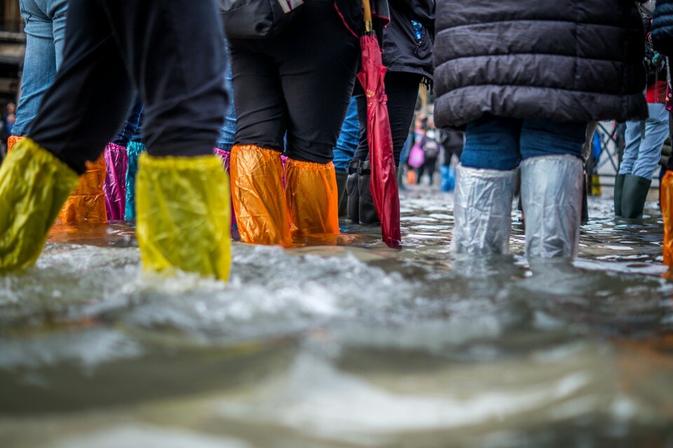 Erweitern Sie Ihren Versicherungsschutz um Elementargefahren. | © Jonathan Ford/Unsplash Auf dem Bild sieht man die Beiden von Menschen in bunten Gummiestiefeln durch eine überflutete Straße gehen. | © Jonathan Ford/Unsplash