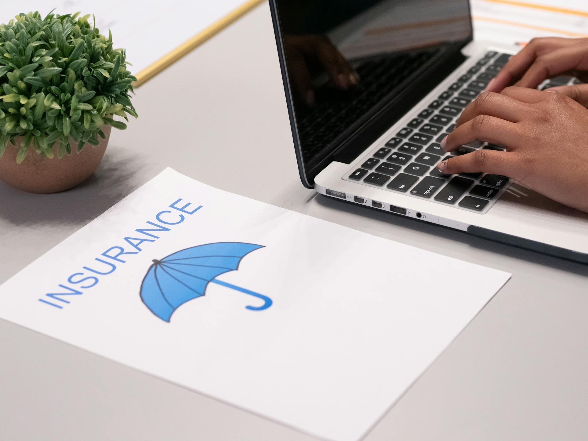 Auf dem Bild sehen Sie eine Laptop und ein Papier, auf dem ein Regenschirm und das Wort insurance abgebildet sind. | © Kindel Media/Pexels.com