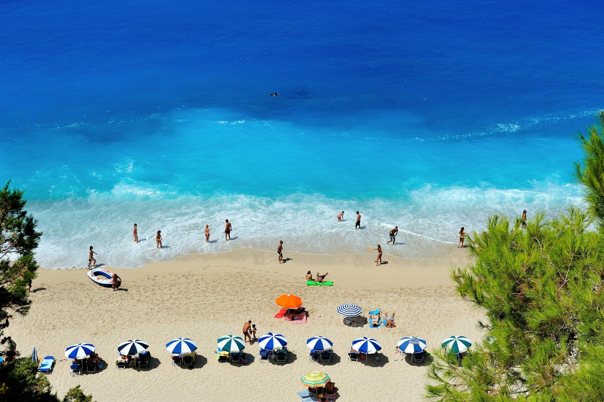 Urlaub am Meer oder im Freibad: Diebstahlschutz und Versicherung | © Nikos Zacharoulis / Unsplash Auf dem Bild sieht man einen Strand mit mehreren blau-weißen und ein paar bunten Sonnenschirmen und Personen, die mit den Füßen im Wasser stehen. Das Meer hat eine tiefblaue Farbe | © Nikos Zacharoulis / Unsplash