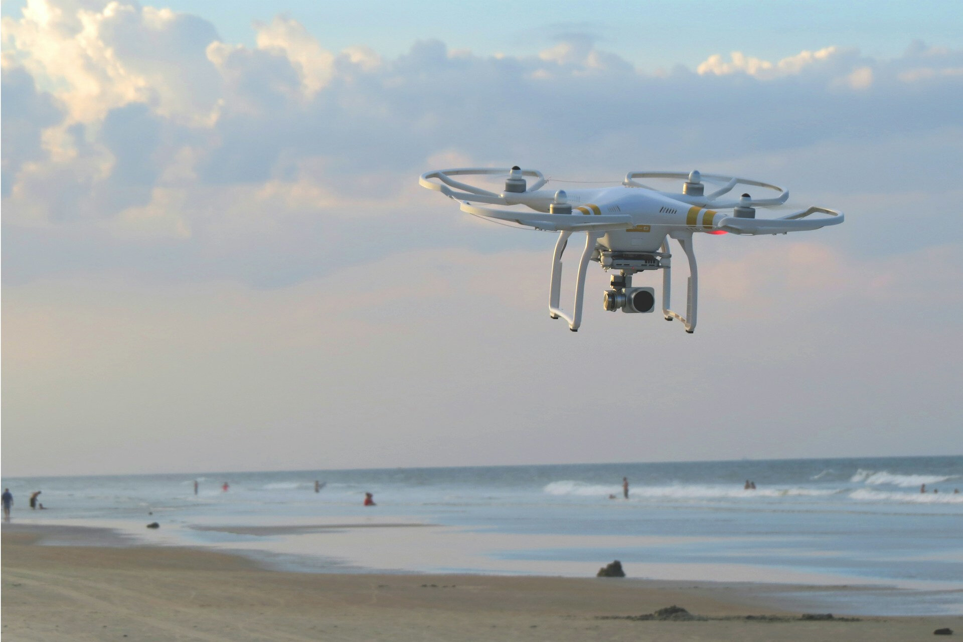 Auf dem Bild sieht man eine Drohne, die am Strand fliegt. Im Hintergrund sieht man das Meer und ein paar Personen | © Matt Pritchard / Unsplash