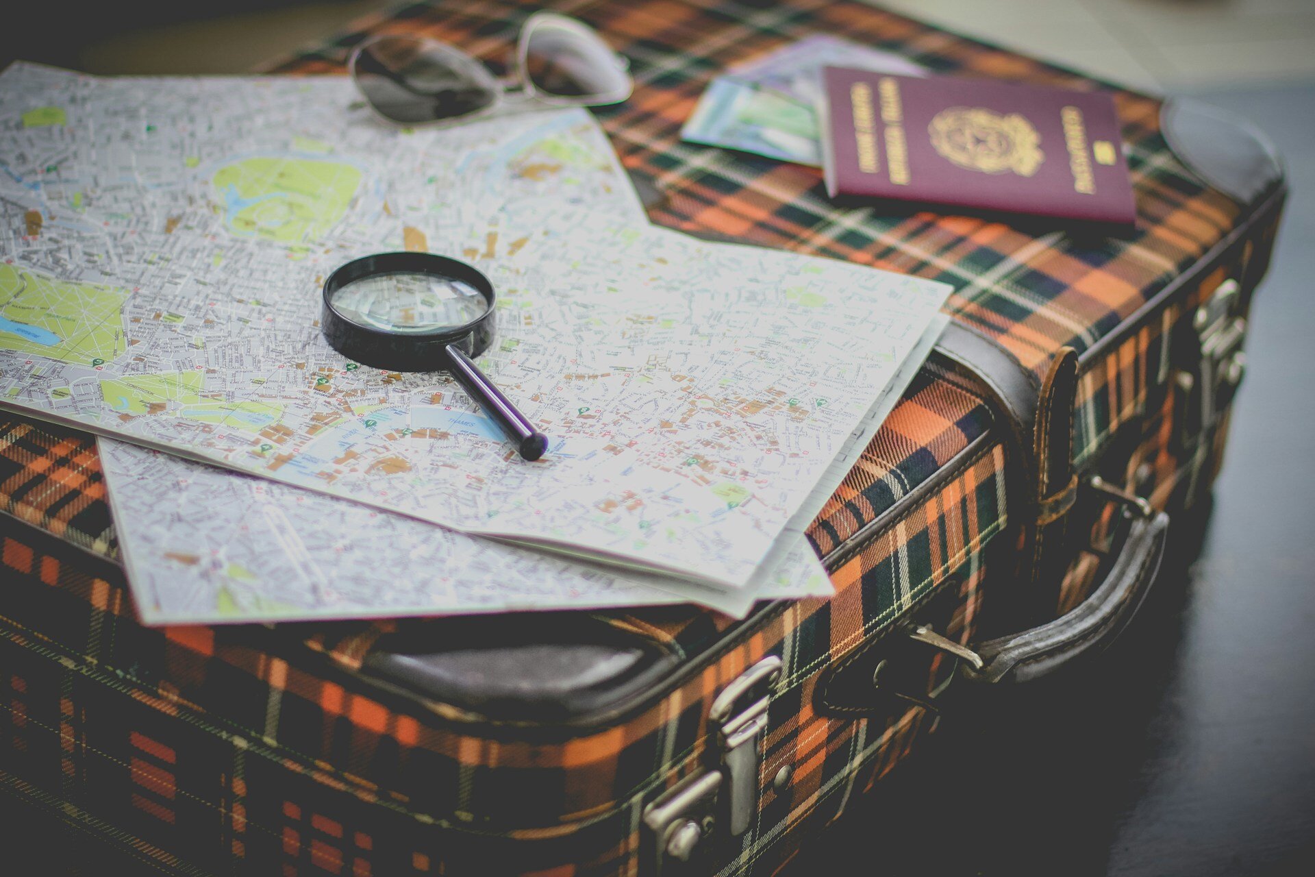 Gut versichert auf Reisen: Tipps zur Auslandsreiseversicherung | © Francesca Tirico / Unsplash Auf dem Bild sieht man den Ausschnitt eines karierten Koffers, auf dem eine Landkarte und eine Lupe liegt. Im Hintergrund kann man noch eine Sonnenbrille und einen Reisepass erkennen | © Francesca Tirico / Unsplash