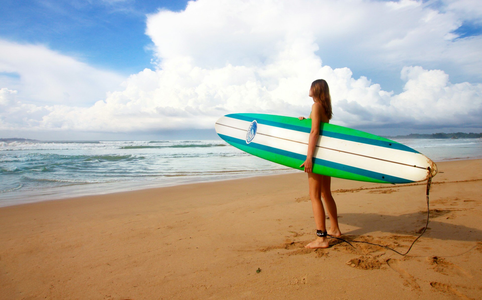 Wichtige Reiseversicherungen: Was du wissen musst | © Filipp Nekhaev/Unsplash.com Auf dem Bild steht eine Frau-sin der Nähe des Meeres und hält ein weiß-blau-grünes Surfbrett. Der Himmel ist blau. | © Filipp Nekhaev/Unsplash.com