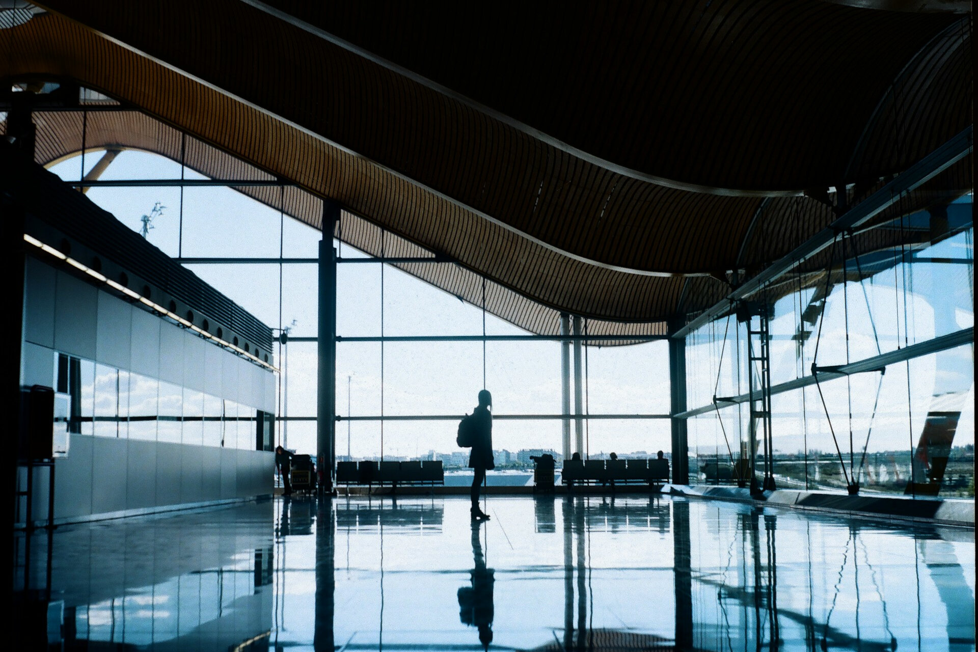Silhouette einer Person, die durch einen Flughafen geht, umgeben von Reisenden und Gepäckwagen. | © ilya / Unsplash