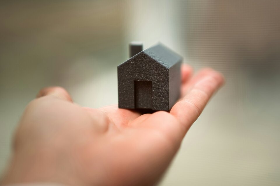 Das Bild zeigt eine Handfläche, auf der ein kleines schwarzes Haus steht. | © Jakub Żerdzicki / Unsplash
