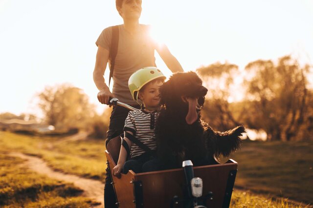  Ein Mann und ein Kind fahren auf einem Lastenrad, begleitet von einem Hund.